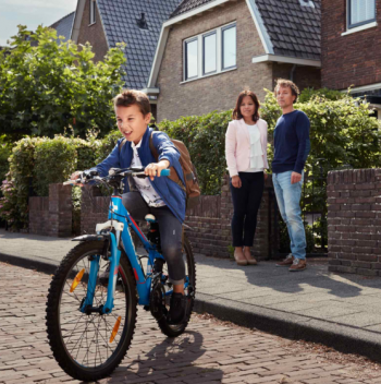 Twee ouders staan voor het huis te kijken naar hun zoontje die weg fietst op zijn blauwe fiets
