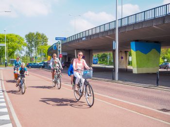 Twee moeders en een vader fietsen met hun kinderen achterop langs onder de brug.